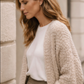 Woman wearing a beige cardigan over a white shirt and khaki pants against a light-colored wall.