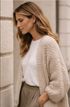 Woman wearing a beige cardigan over a white shirt and khaki pants against a light-colored wall.