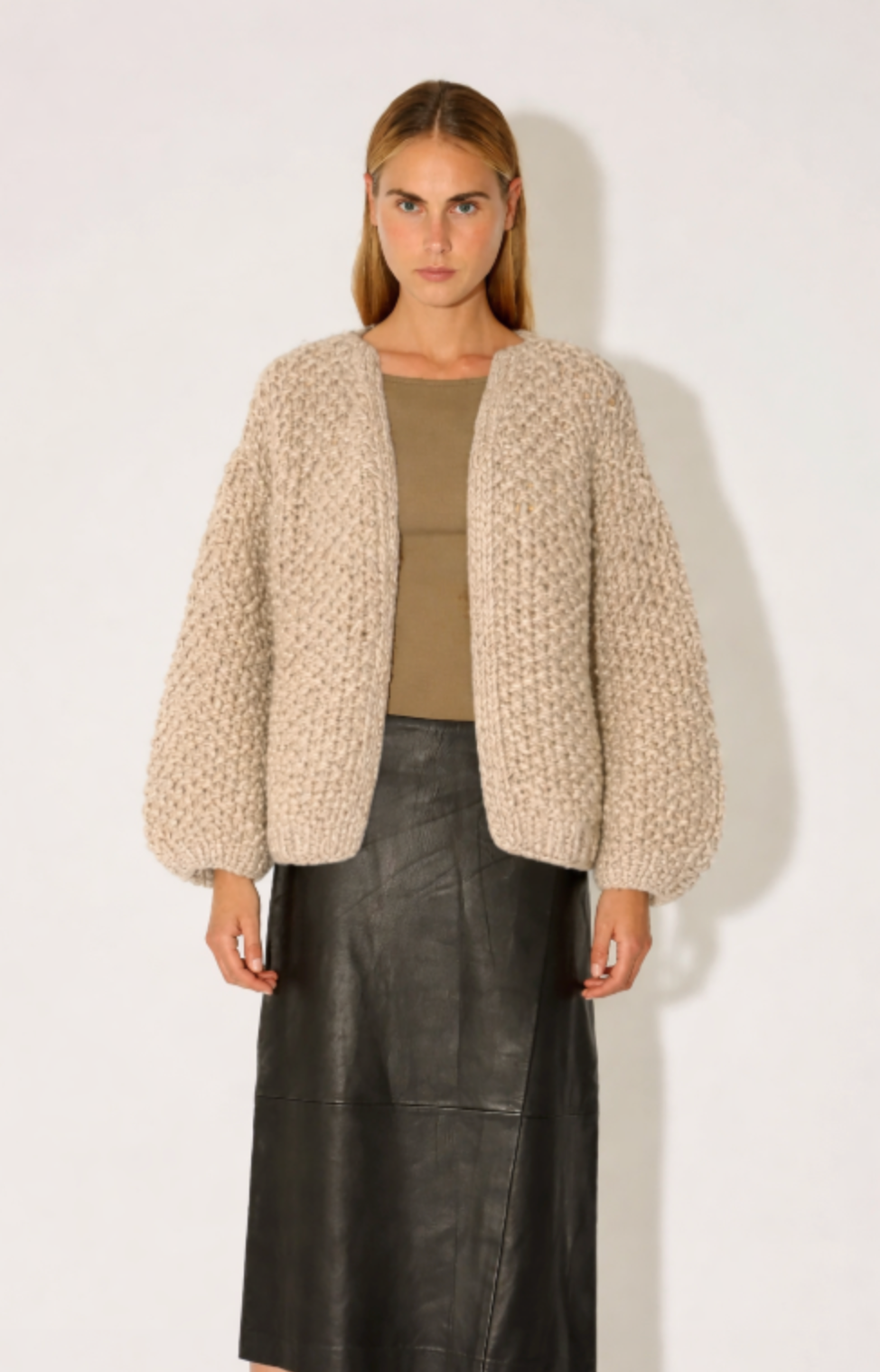Woman wearing a beige textured cardigan over a green top and black leather skirt on a white background