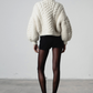 Person wearing a white chunky knit sweater and black shorts with fishnet tights on a plain background