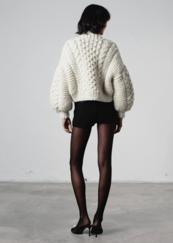 Person wearing a white chunky knit sweater and black shorts with fishnet tights on a plain background