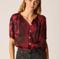 Woman wearing a red blouse with black patterns and dark shorts on a beige background