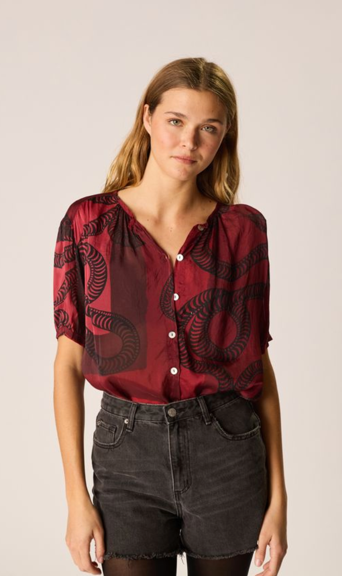 Woman wearing a red blouse with black patterns and dark shorts on a beige background